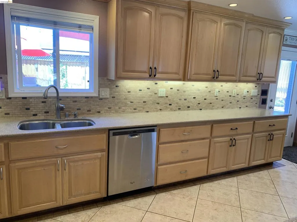 Kitchen featuring light wood cabinetry, a double basin stainless steel sink with a gooseneck faucet, light-toned countertops, a mosaic tile backsplash, and a stainless steel dishwasher