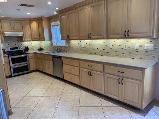 Spacious kitchen featuring light wood-finish cabinetry, a full suite of stainless steel appliances including a double oven range and dishwasher, an undermount sink with a gooseneck faucet, light-toned countertops, and a mosaic tile backsplash