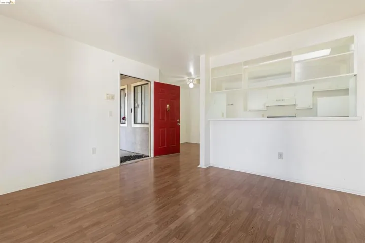 Open-concept living area featuring wood-finish flooring, a vibrant red entry door, a kitchen pass-through with open shelving, an integrated range hood, and a ceiling fan