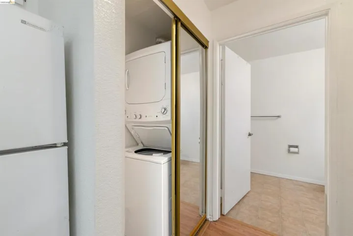 Stackable washer and dryer unit within a mirrored closet, complemented by a white refrigerator and wood-finish flooring