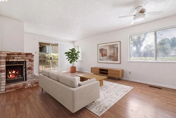 Living area featuring wood-finish flooring, a brick fireplace, a ceiling fan, and large windows with blinds