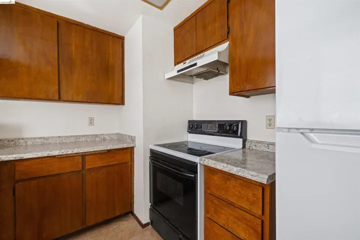Compact kitchen featuring wood-finish cabinetry, light-toned countertops, and a white electric range with an overhead exhaust fan