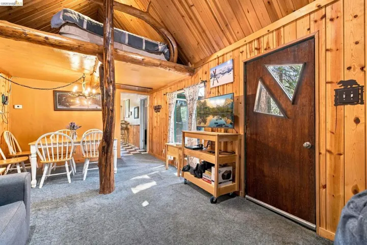 Rustic wood-paneled interior featuring a lofted sleeping area, exposed log support beams, a dining area with pendant lighting, and a unique entry door with triangular window inserts