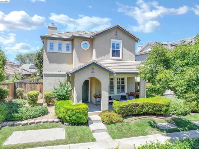 Two-story stucco exterior featuring a covered entryway, tiled roof, decorative circular window, and mature front landscaping with manicured shrubs
