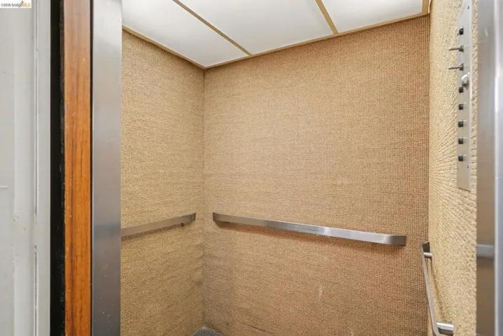Elevator interior featuring textured wall coverings, stainless steel handrails, a metal control panel, and a wood-finish door frame