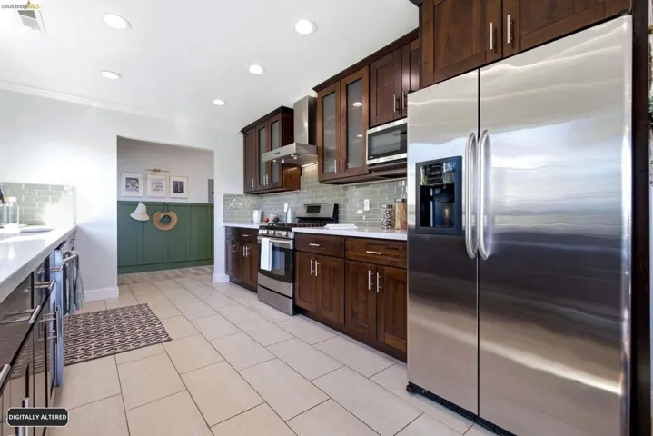 Kitchen featuring wood cabinetry, stainless steel appliances, and light-toned tile flooring. Pantry and area leading to backyard and lower level