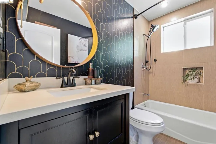 Hallway Bathroom featuring a dark wood-finish vanity with a light-toned countertop, a gold-framed circular mirror, a built-in bathtub with a tile surround, a shower system with matte black hardware, and an integrated shower niche