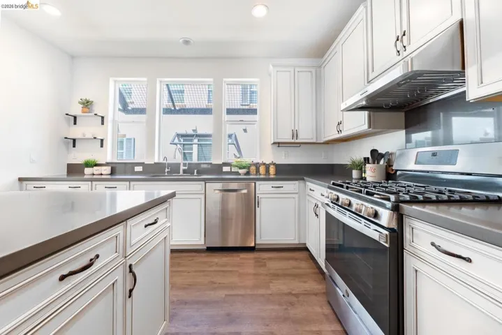 Modern kitchen featuring white shaker cabinetry, stainless steel appliances, dark countertops, a gas range with an overhead vent, and wood-finish flooring