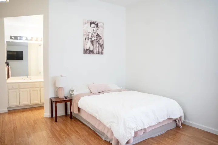 Wood-finish flooring throughout, bright white walls, and an attached bathroom featuring a vanity with a mirror and overhead lighting