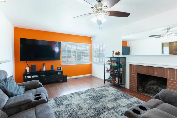 Living area featuring wood-finish flooring, a brick-surround fireplace, and a ceiling fan with integrated lighting