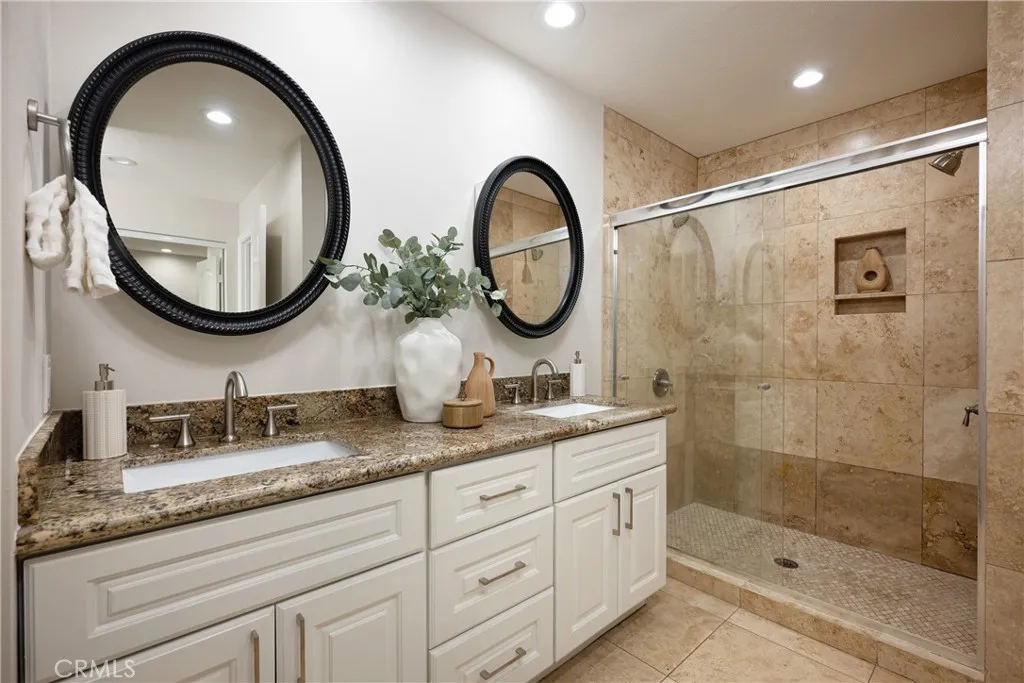 The beautifully remodeled primary bathroom features dual vanities, granite countertops, white cabinetry, travertine finishes, and a spacious walk-in shower.