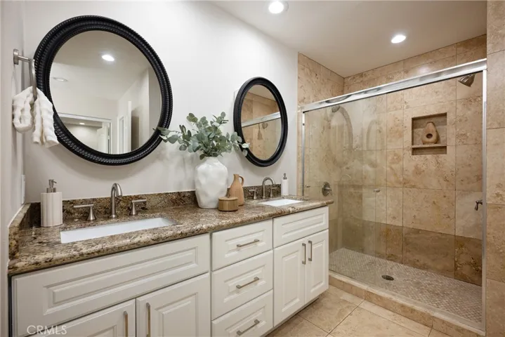 The beautifully remodeled primary bathroom features dual vanities, granite countertops, white cabinetry, travertine finishes, and a spacious walk-in shower.