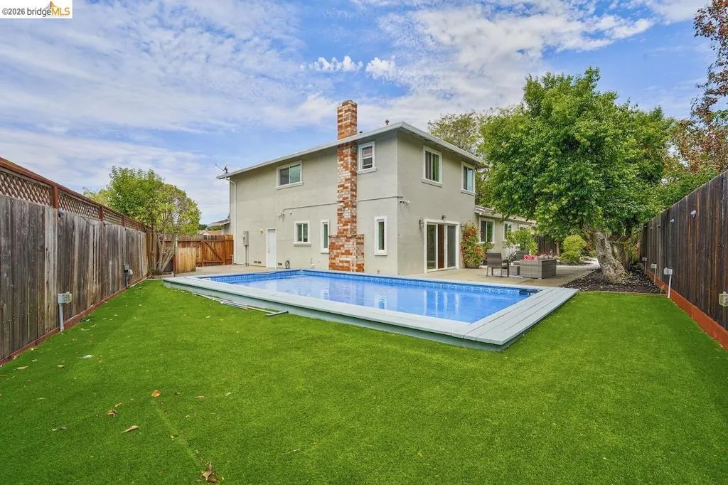 Backyard featuring an in-ground swimming pool with light blue tiling, a surrounding artificial turf lawn, and a light-colored stucco exterior with a brick chimney