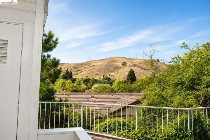 Balcony featuring white railings and a partial louvered door, overlooking a terraced landscape with mature trees and distant hills