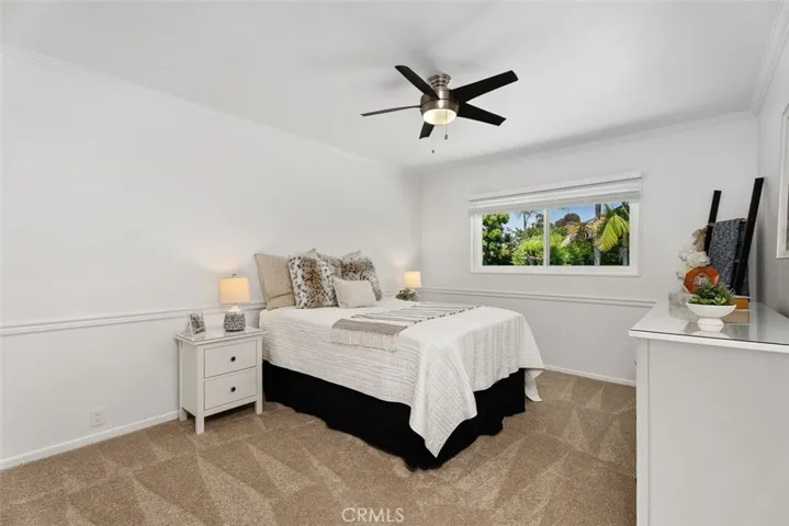 This light and bright bedroom features crisp white walls, neutral carpeting, a classic ceiling fan, and a wide window overlooking mature trees.
