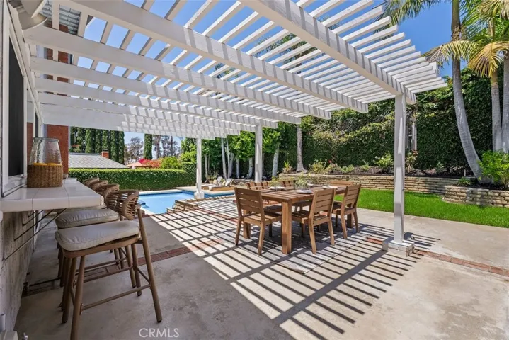 The backyard boasts a massive white wood-slat pergola that provides comfortable shade for an outdoor dining table and built-in bar.