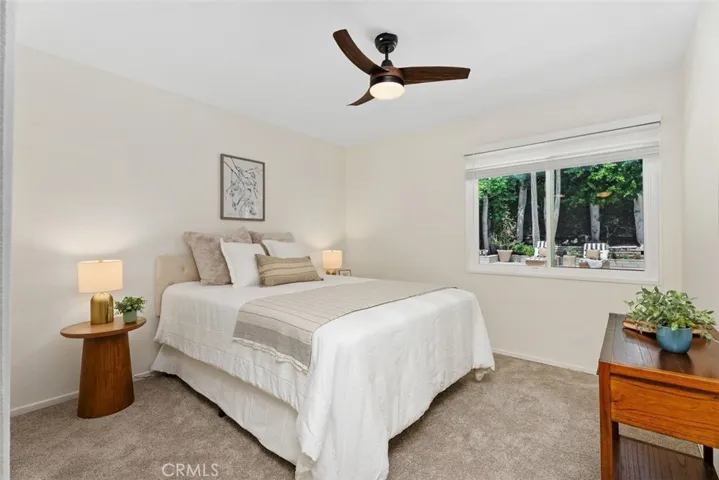 This main-floor bedroom features new carpeting, a modern ceiling fan, and a large window looking out to the lush backyard greenery.