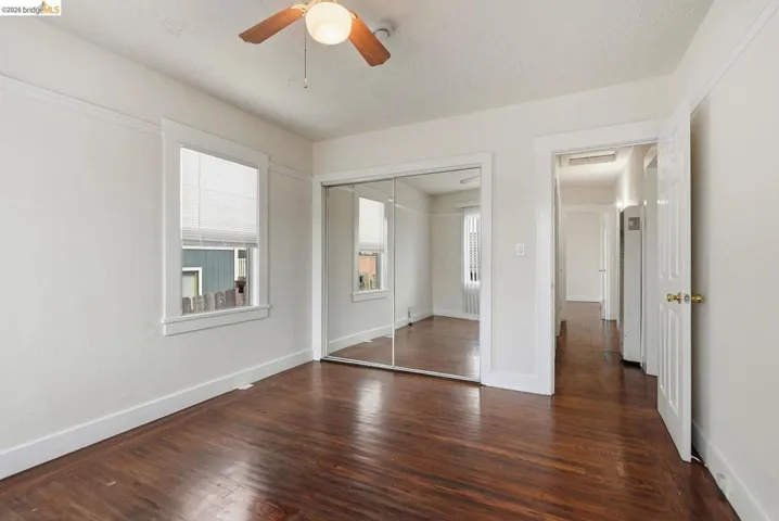 Wood-finish flooring throughout, featuring a ceiling fan with integrated lighting, mirrored sliding closet doors, and a window with blinds