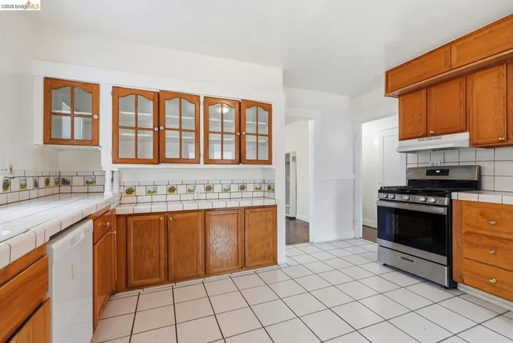 Kitchen featuring white square tile flooring, wood cabinetry with glass-front upper cabinets, a stainless steel range with overhead ventilation, and tiled countertops with a decorative backsplash