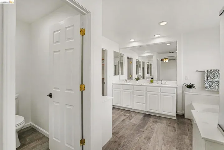 Bathroom featuring wood-finish flooring, a double vanity with white cabinetry, a large wall-mounted mirror, recessed lighting, and a built-in tub with a white surround