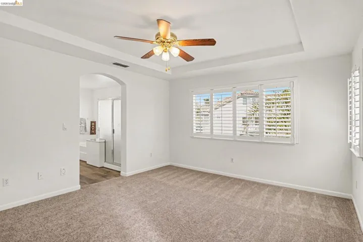 Spacious room featuring tray ceilings with a ceiling fan, plush carpet flooring, and white plantation shutters on multiple windows