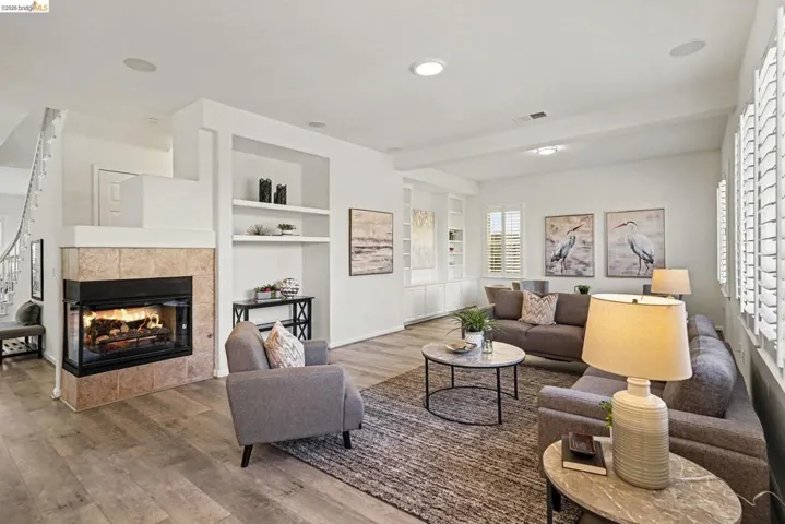 Spacious living area featuring wood-finish flooring, a tiled fireplace with a glass enclosure, white plantation shutters, built-in shelving, and recessed lighting