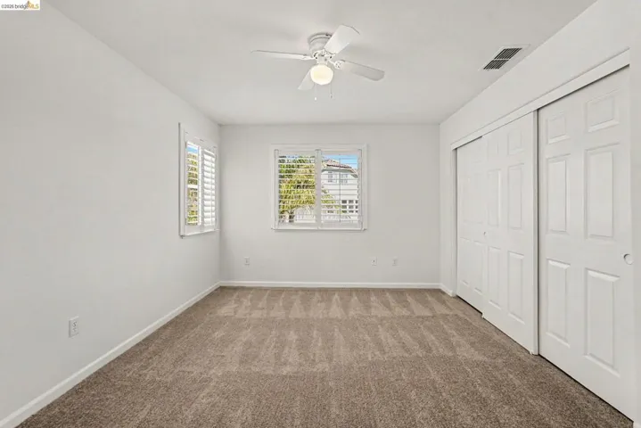 Carpeted interior space featuring neutral wall paint, two windows with plantation shutters, a ceiling fan with light fixture, and double sliding closet doors