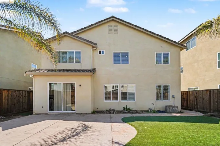 Rear exterior featuring a stucco finish, multiple windows with blinds, a sliding glass door, a concrete patio, and an artificial turf lawn