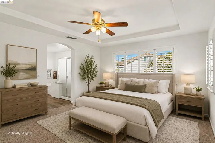 Spacious bedroom featuring a tray ceiling with a ceiling fan, white plantation shutters, and an arched entryway to an en-suite bathroom with a glass-enclosed shower