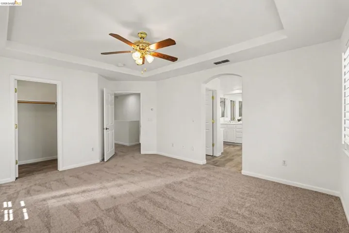 Spacious room featuring tray ceilings with a wood-finish ceiling fan, plush carpet flooring, and white plantation shutters