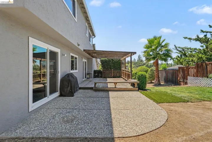 Spacious outdoor area featuring an exposed aggregate concrete patio, a wooden deck with a pergola, a lawn area, mature palm tree, and wooden fencing