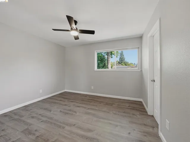 Interior room featuring wood-finish flooring, light gray walls, a ceiling fan with integrated lighting, a white-framed window, and a white door with a silver knob