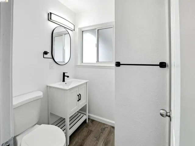 White vanity with integrated sink, black matte faucet, oval mirror, and contemporary light bar over wood-finish flooring