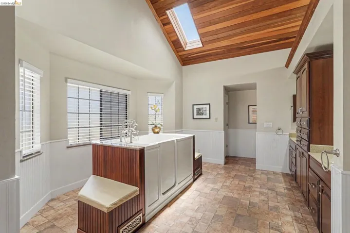 Spacious bathroom featuring a vaulted wood-finish ceiling with a skylight, a large walk-in tub with wood-panel detailing, two window banks with blinds, extensive vanity cabinetry with dual sinks, and tile flooring