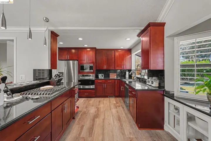 Kitchen featuring warm-toned cabinetry, stainless steel appliances, dark countertops, wood-finish flooring, and a window with white louvered shutters
