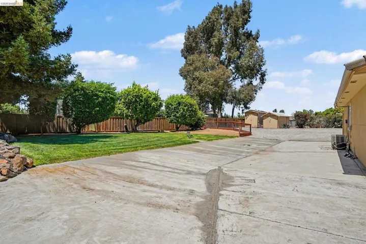 Expansive concrete driveway featuring a grassy area with mature trees, a wood fence, and a wooden ramp