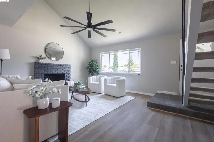 Vaulted ceiling living area featuring a modern black ceiling fan, a painted brick fireplace, large windows, wood-finish flooring, and a tiled entry