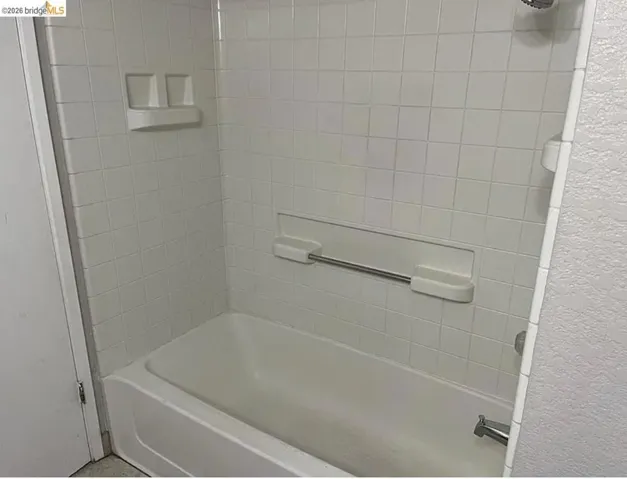 Bathtub and shower combination featuring white tiled walls, an integrated soap dish, and a stainless steel shower head and faucet