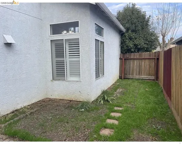 Exterior corner featuring stucco siding, multiple windows with louvered shutters, a stepping stone path, and a wooden privacy fence