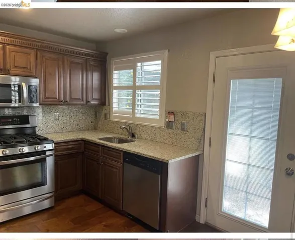 Kitchen featuring wood-finish flooring, stainless steel appliances, granite countertops with a matching backsplash, rich wood cabinetry, and a window with plantation shutters