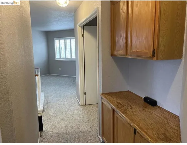 Hallway storage cabinetry with wood-finish doors and countertop, featuring overhead flush-mount lighting, and light-toned carpet flooring leading to a room with window shutters