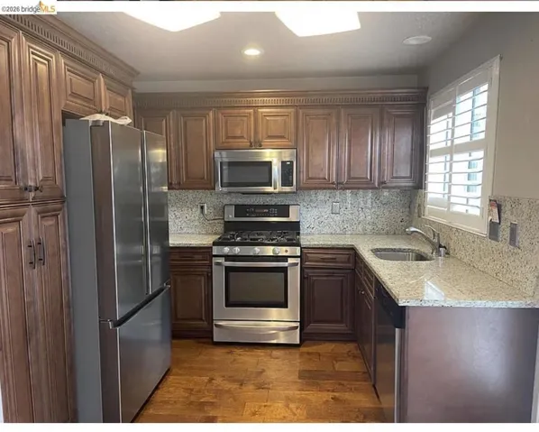 Kitchen featuring wood-finish flooring, dark wood cabinetry with crown molding, stainless steel appliances, speckled countertops, and a matching backsplash