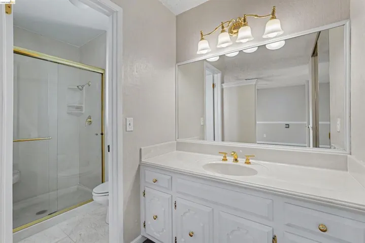 Vanity with dual-handle faucet and integrated sink, large wall-mounted mirror, four-light vanity fixture, white cabinetry with gold-tone hardware, and a glass-enclosed shower