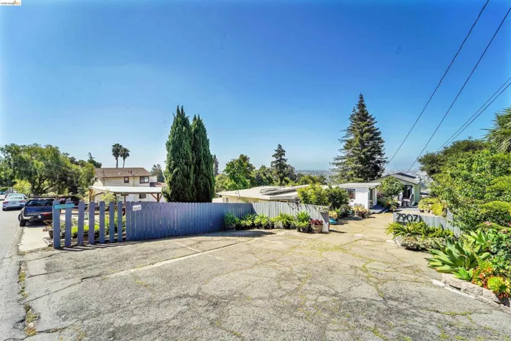 Paved driveway entry with a blue picket fence, bordered by mature trees and varied landscaping