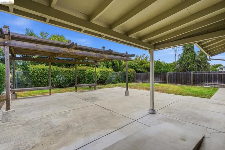 Expansive concrete patio area with a wooden pergola structure, covered patio extension, lush green lawn, and a privacy fence