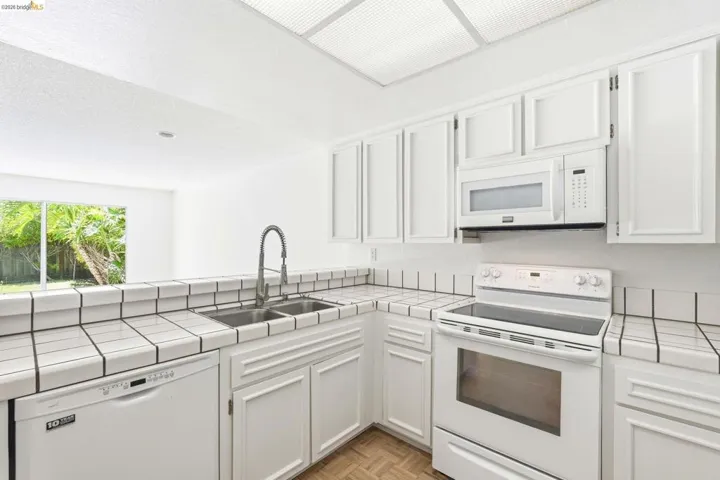 Kitchen featuring white cabinetry and appliances, tile countertops with a tile backsplash, a double basin stainless steel sink with a gooseneck faucet, and wood-finish flooring