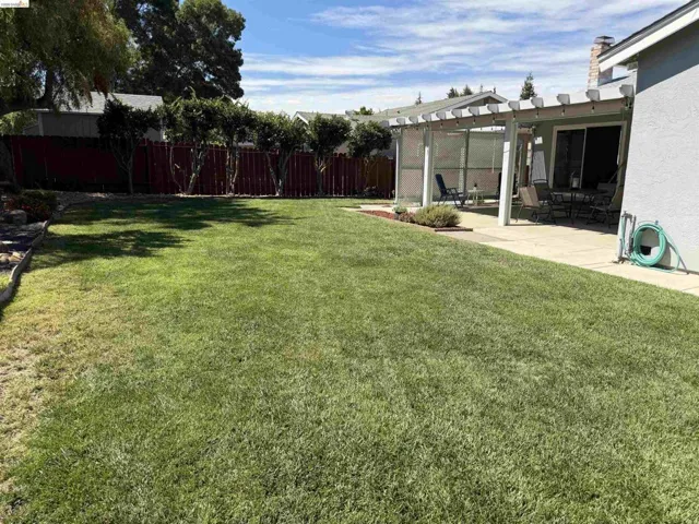 Expansive lawn area featuring a concrete patio with a white pergola, bordered by a red privacy fence and mature trees