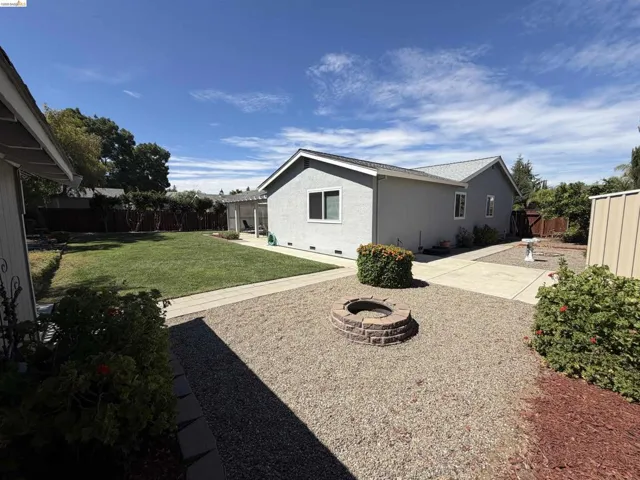 Expansive backyard featuring a manicured lawn, concrete patio, a gravel area with a built-in fire pit, mature landscaping, and a detached structure