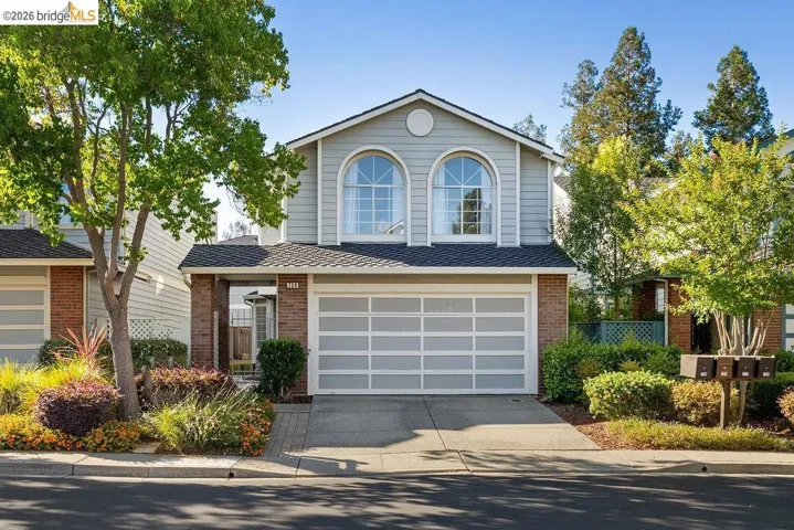 Two-story residence featuring light gray siding with brick accents, an attached garage, two arched windows, and mature landscaping