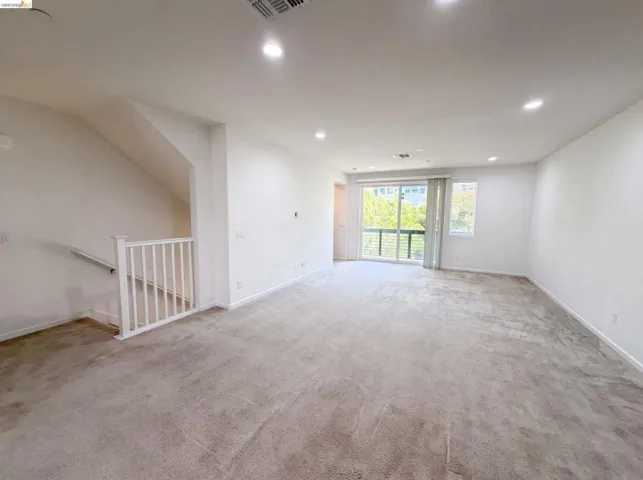Spacious carpeted room featuring recessed lighting, white painted walls, a white staircase railing, and a glass sliding door leading to a balcony